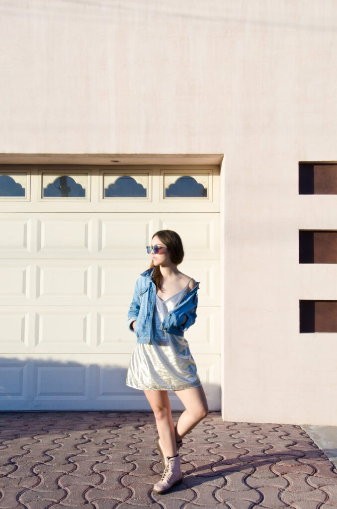 Laura Adriana photo shoot styled in a shimmering dress denim jacket and pink boots poses confidently against a pastel pink wall with a white garage door featuring scalloped windows.